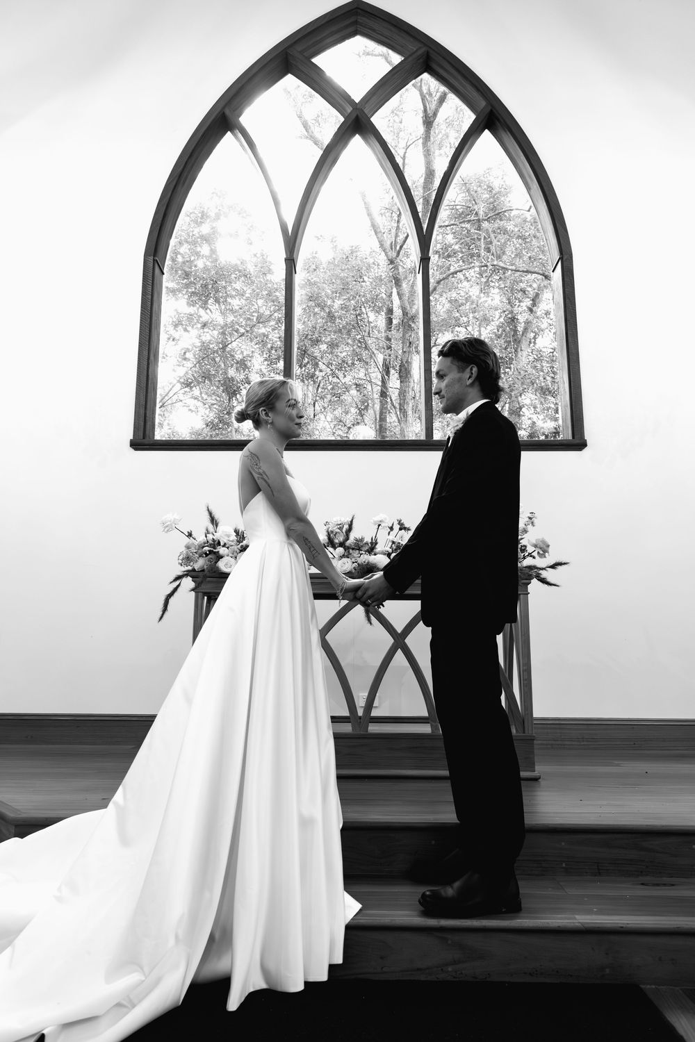 A classic black and white photo of a couple at the altar with a beautiful arched chapel window behind them.