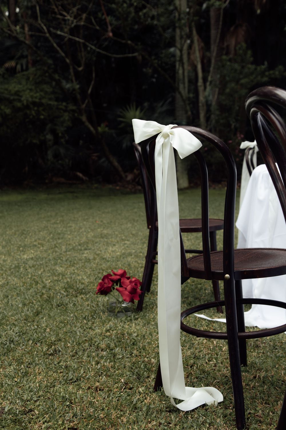 Row of dark wooden chairs decorated with white ribbons and red floral accents set up for an outdoor garden ceremony.