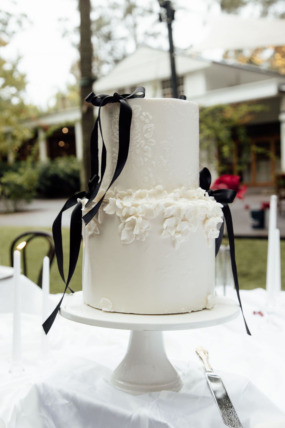 A round white wedding cake decorated with intricate lace details and black ribbon on an elegant white pedestal stand.
