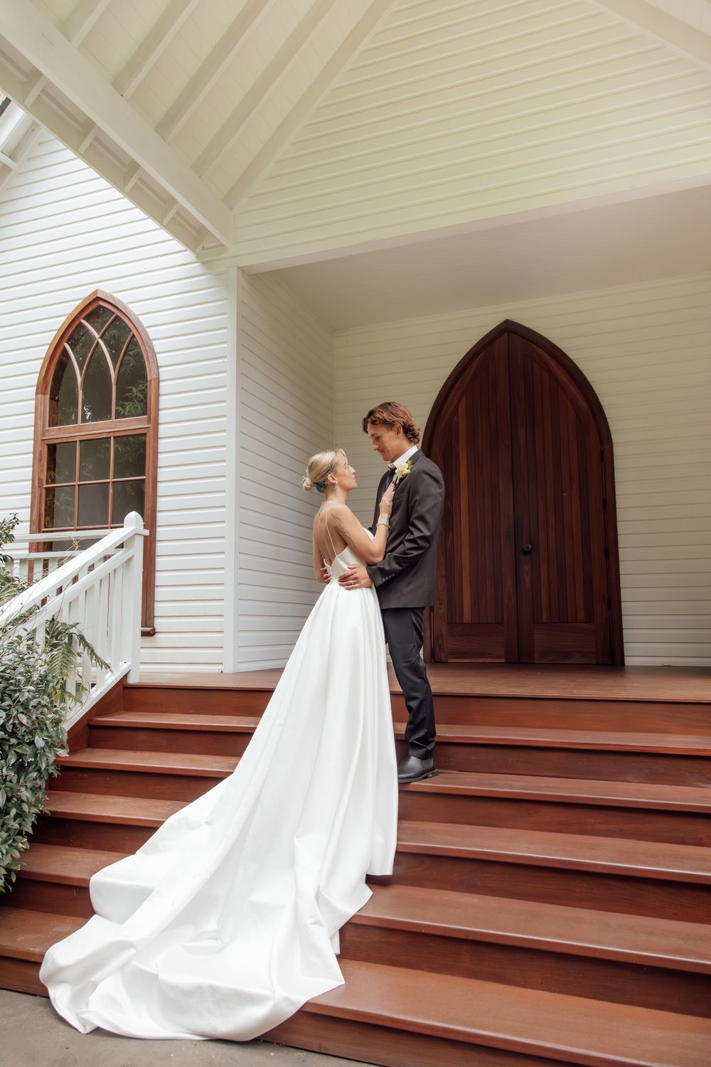 A couple stands romantically on the steps of a white chapel with gothic windows and a wooden door.