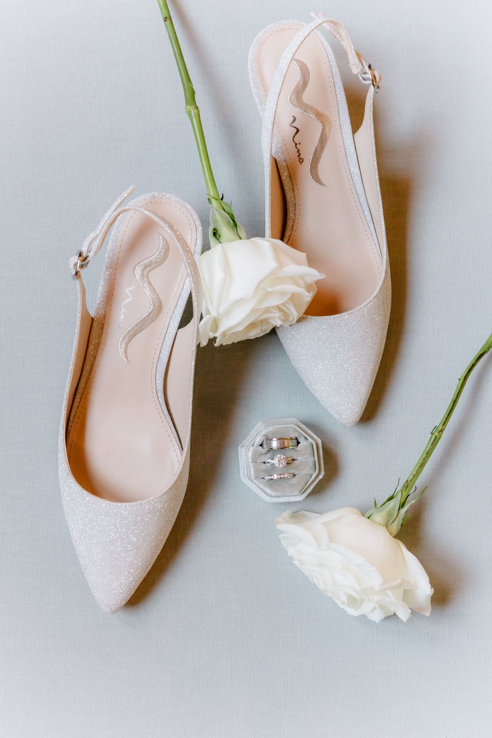 Elegant white bridal shoes arranged with delicate white roses against a light gray background.