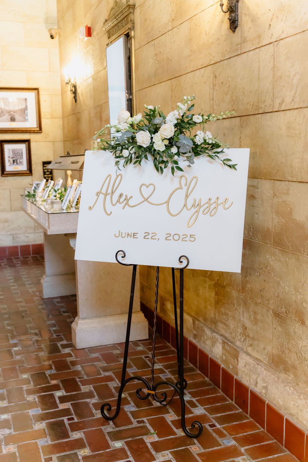 Elegant wedding signage displayed on iron stands with white floral accents along a rustic brick floor hallway.
