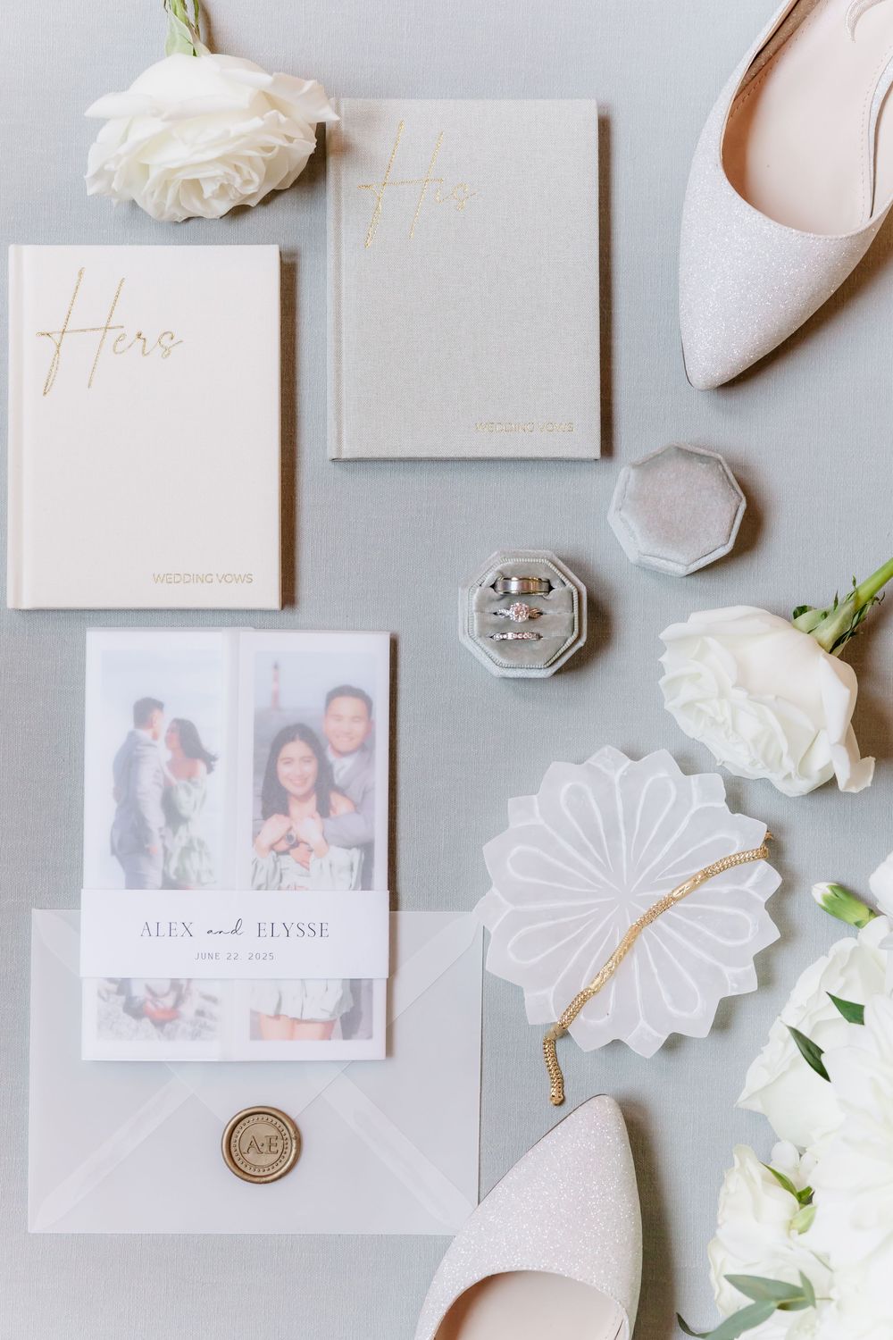 Elegant wedding stationery flatlay with white invitations, gold wax seals, ivory roses and blush pink shoes on gray background.