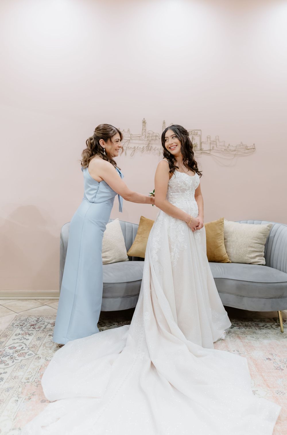 Two people in light blue and white dresses share a tender moment while getting ready for a wedding celebration.
