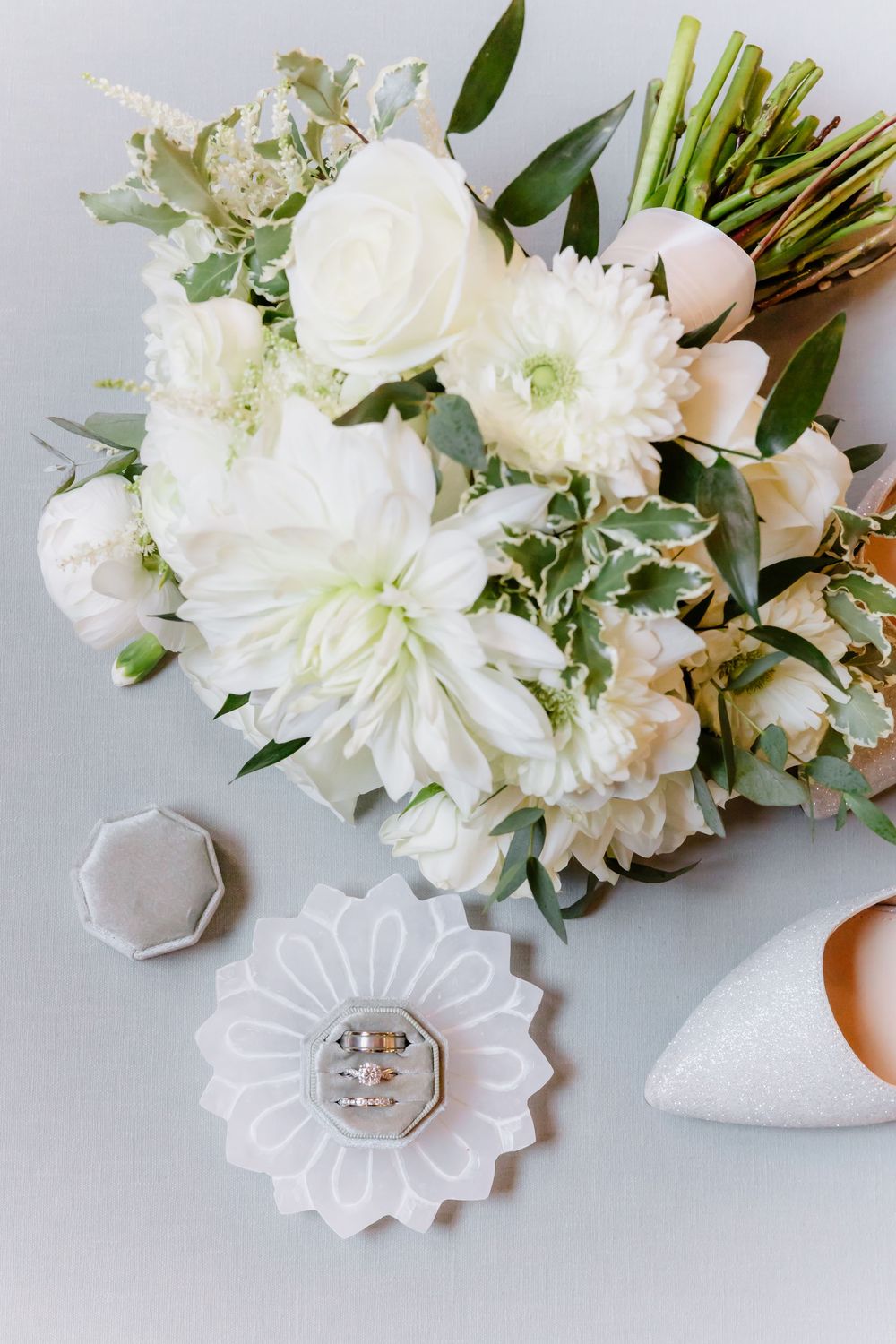 Elegant white wedding bouquet with roses and peonies arranged beside decorative ring dishes on light gray surface.