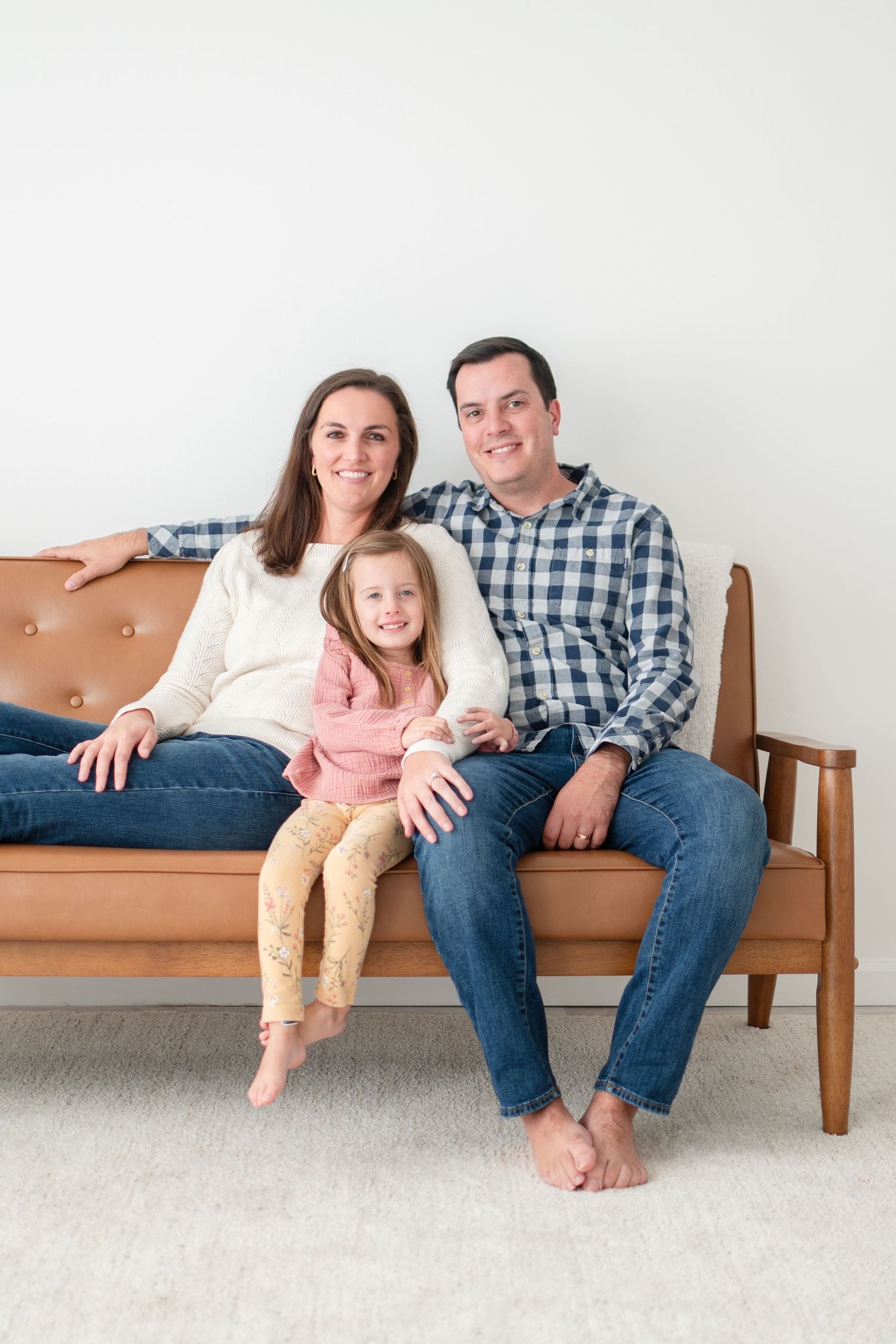 A family sits together on a brown wooden bench against a light colored wall in coordinating casual outfits.