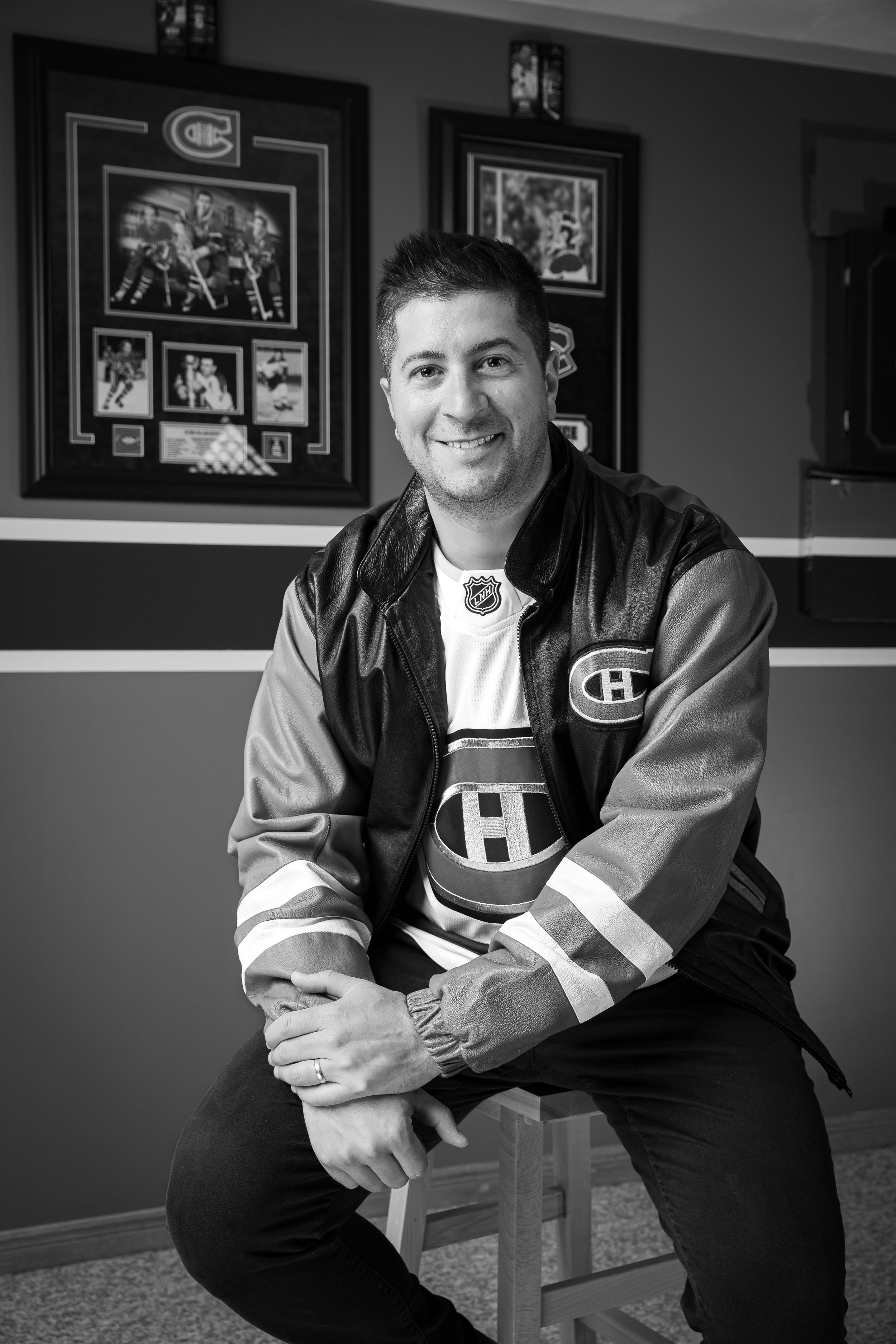 A black and white photo of someone wearing a Montreal Canadiens NHL jacket sitting on a stool with framed sports memorabilia behind.