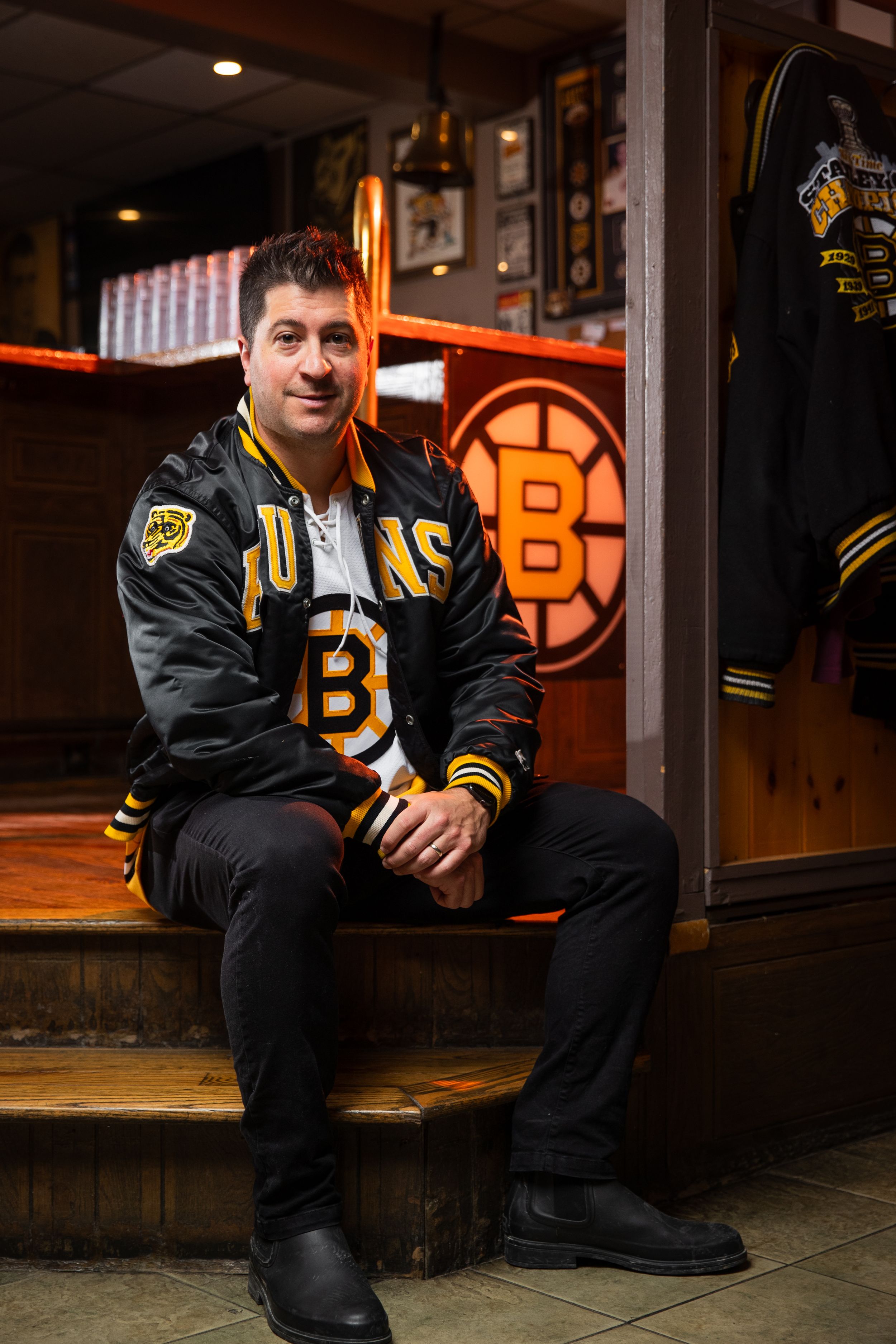 A person wearing a Boston Bruins jacket sits on wooden steps inside a hockey themed venue with team logos on display.