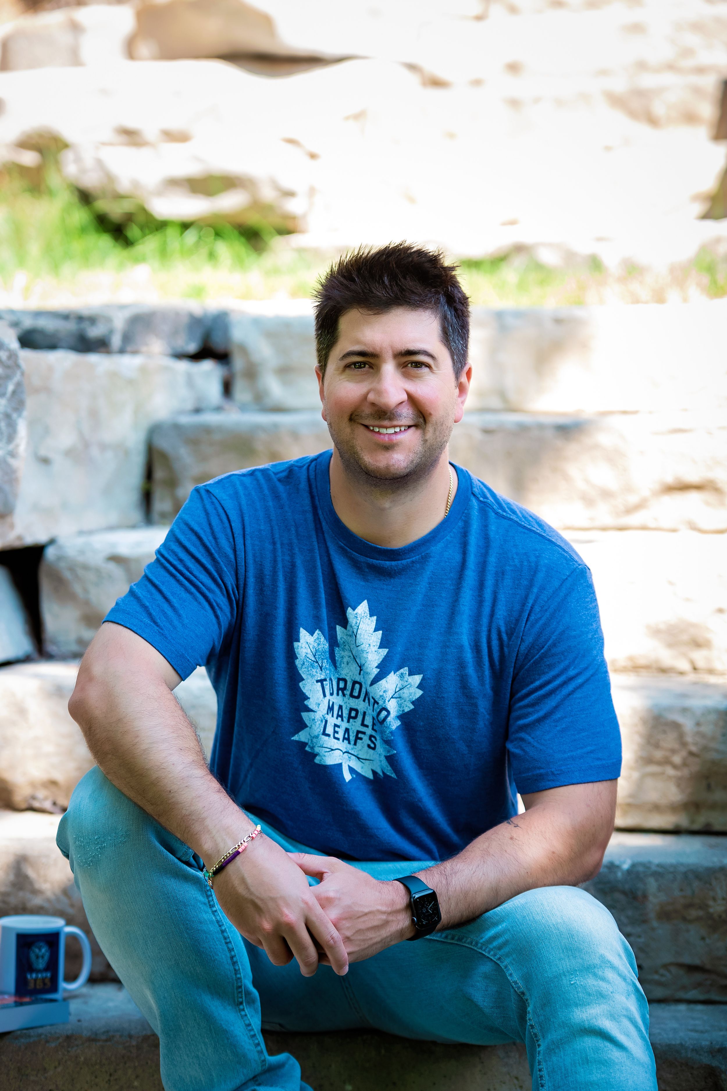 A person wearing a blue Toronto Maple Leafs t-shirt and jeans sits casually on stone steps outdoors.