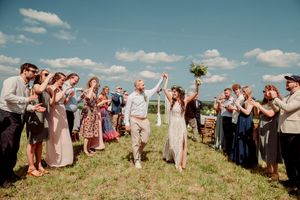 Joyful newlyweds walk through cheering guests outdoors on a sunny day, bride holding bouquet aloft in a field.