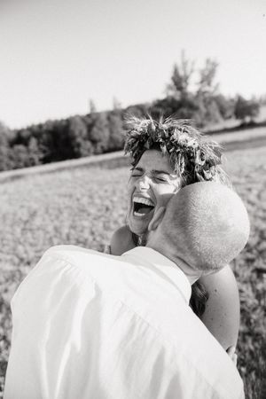 Black and white photo of a joyful bride wearing a flower crown laughing while being embraced by a groom outdoors.