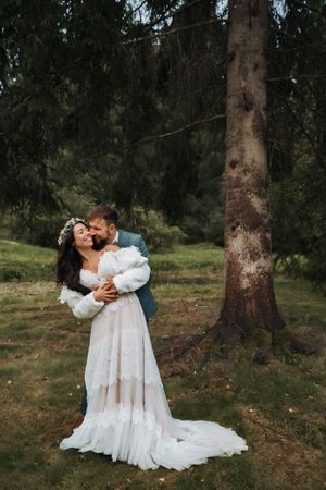 Romantic embrace between couple in flowing white dress among tall forest trees.