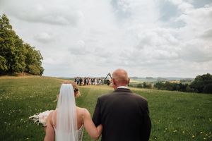 Bride and groom walking hand in hand toward outdoor wedding ceremony on lush green hillside under cloudy sky.