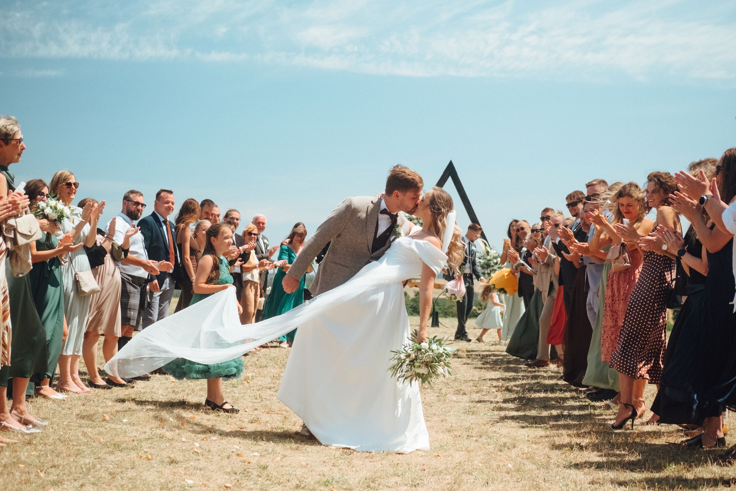 Bride and groom sharing a romantic dip kiss at outdoor wedding ceremony with guests applauding under blue sky.