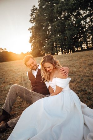 Bride and groom sitting together in golden sunset light on a grassy hill, sharing a romantic moment in wedding attire.