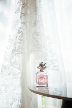 Pink Marc Jacobs perfume bottle with floral cap displayed on a table against sheer white lace curtains.