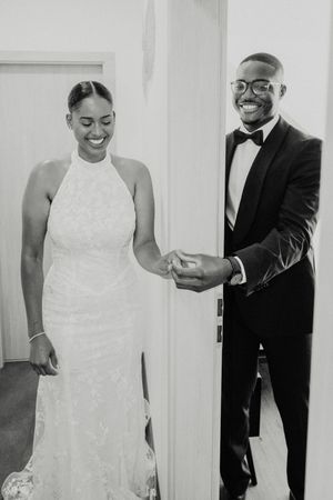 Black and white photo of a smiling bride in halter gown and groom in tuxedo holding hands on their wedding day.