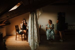 Bride and bridesmaid sit in a rustic attic room with a lace wedding dress hanging nearby in soft dramatic lighting.