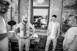 Black and white photo of men in suits socializing outdoors near a rustic stone wall at a casual event.