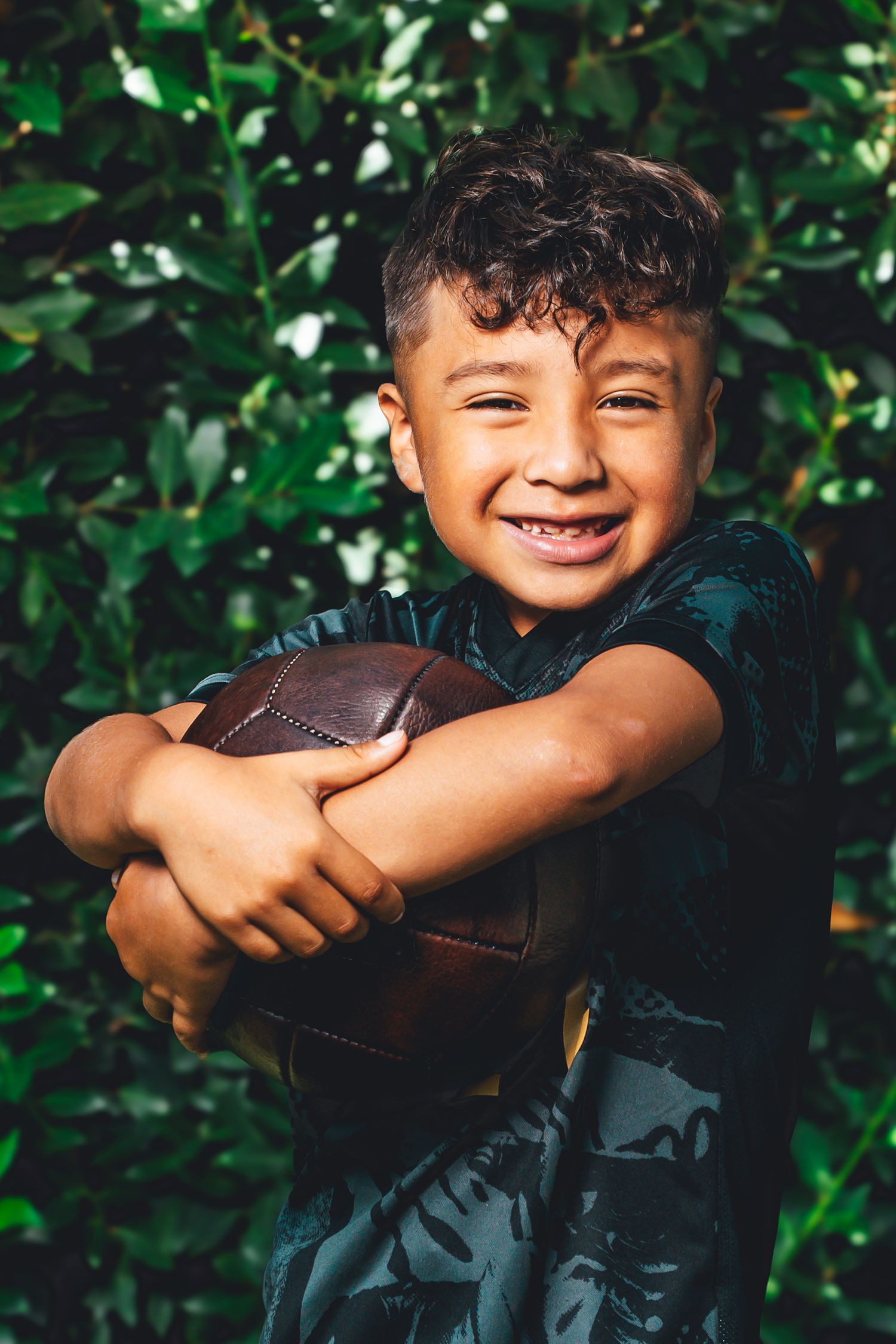 Young child laughing and hugging a soccer ball while surrounded by green foliage in outdoor setting.