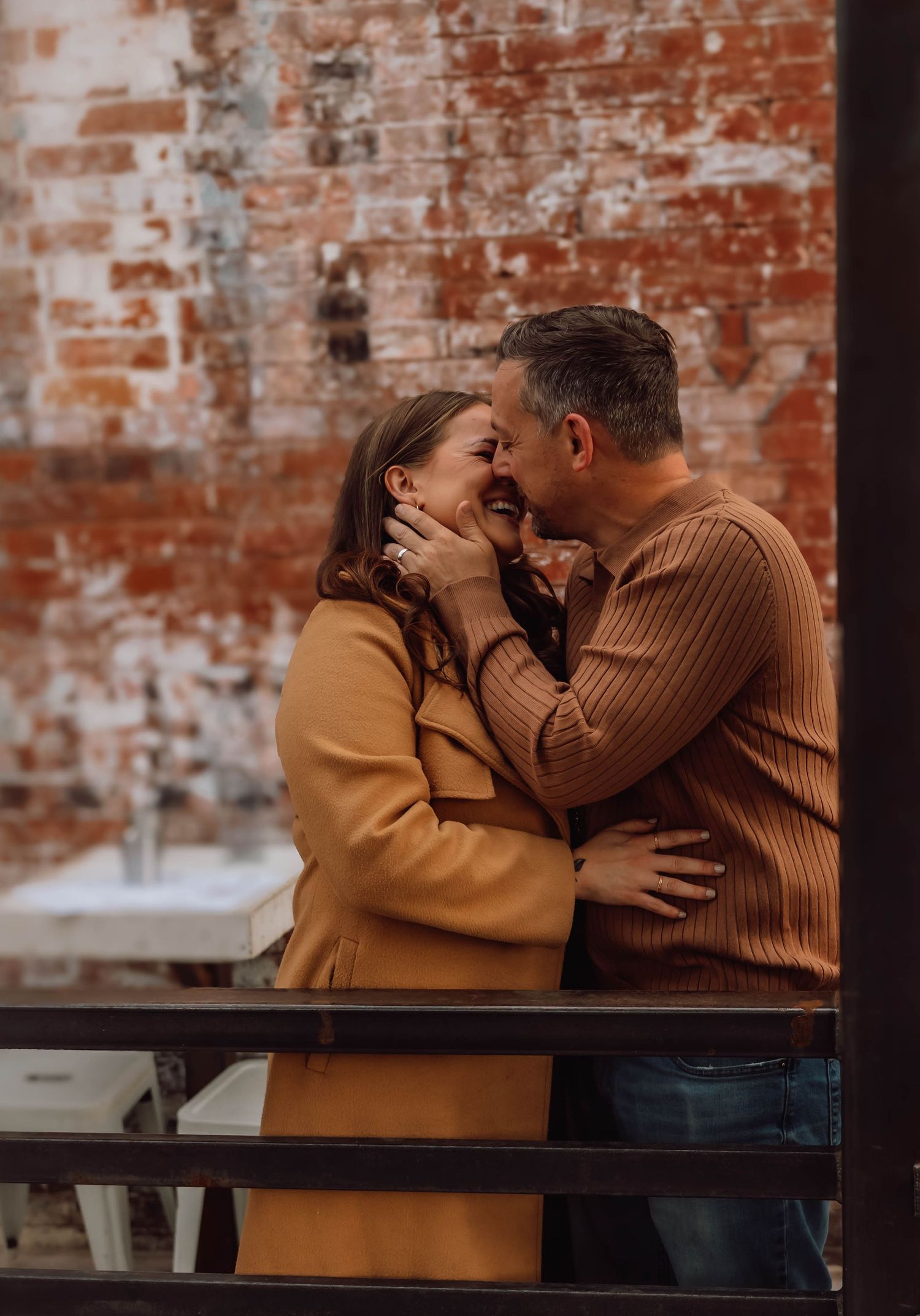 A romantic couple in warm sweaters sharing an intimate moment against a rustic brick wall.