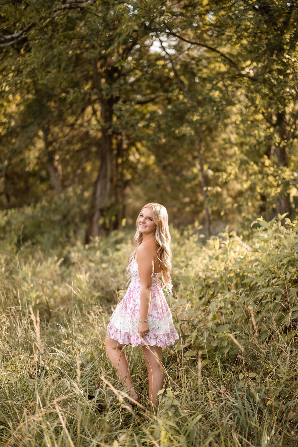 A floral dress stands out against tall grasses in a sunlit field with trees in the background during golden hour.