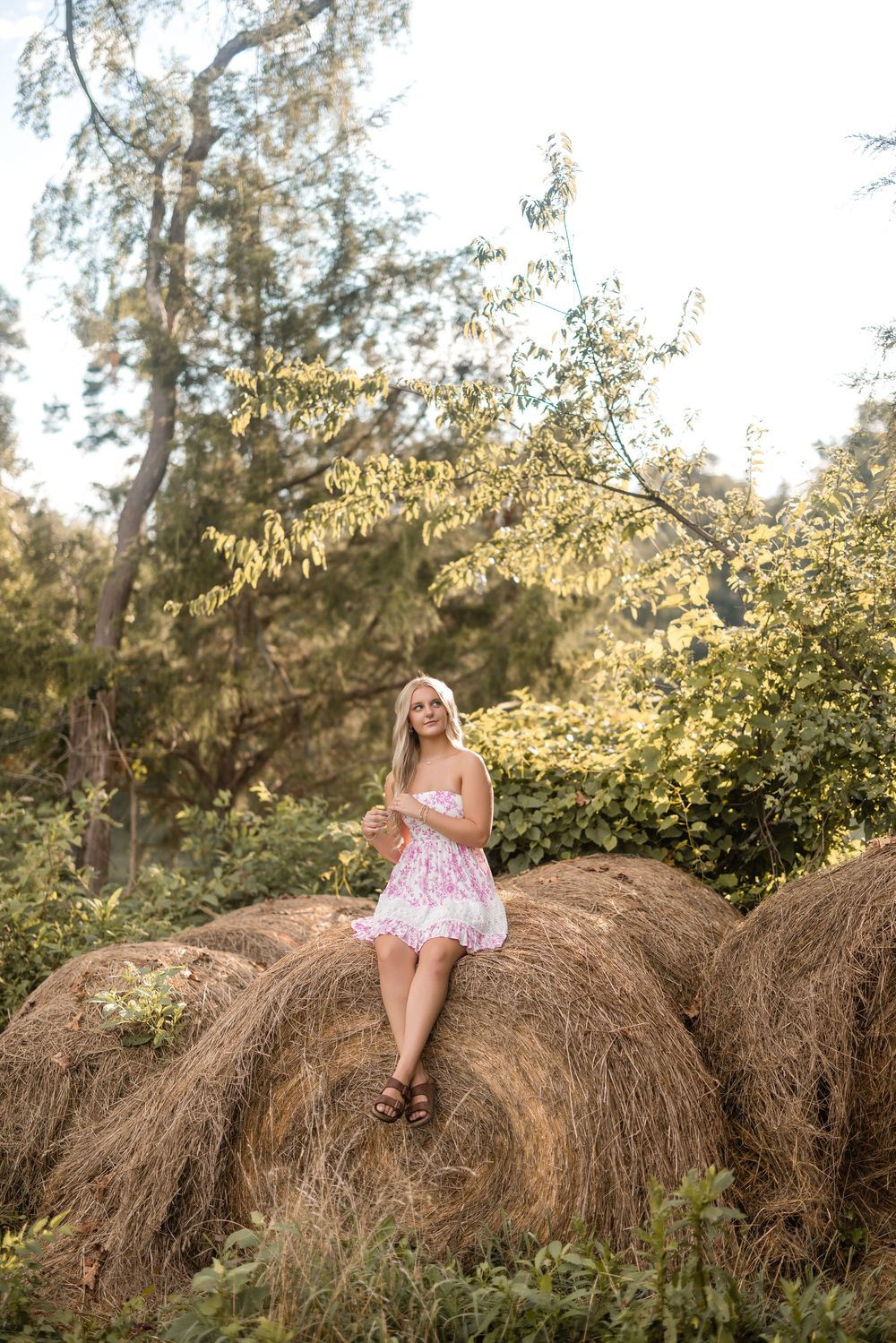 Someone sitting casually on a large hay bale in a rustic outdoor setting during sunset.