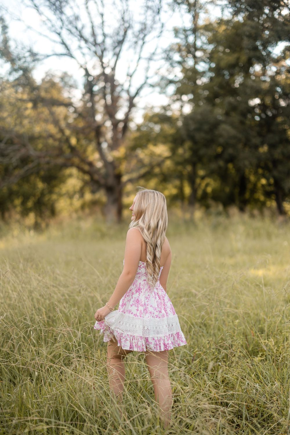 A floral pink dress twirls gracefully in a sunlit meadow with tall grass and trees in the background.