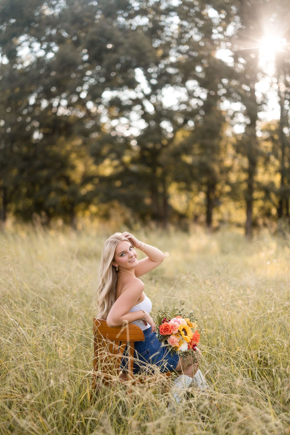 Woman in orange top and jeans lying in golden field during sunset with bouquet of sunflowers.