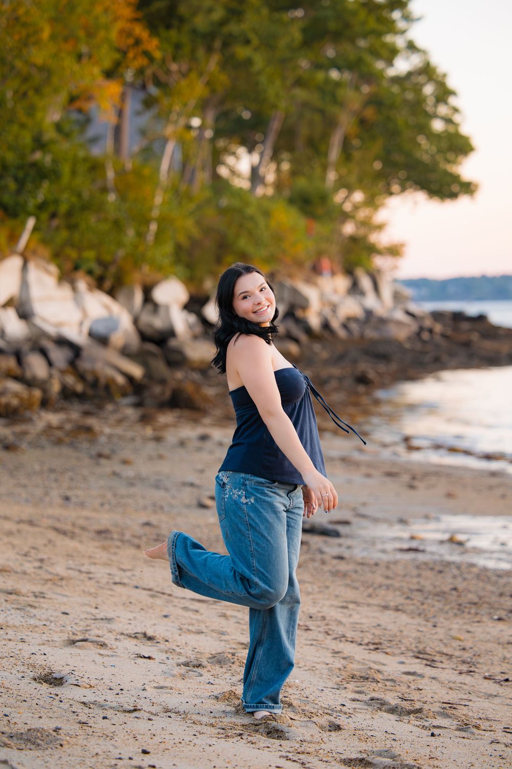 A casual autumn photoshoot shows someone dancing playfully on a beach with trees and rocks in the background.