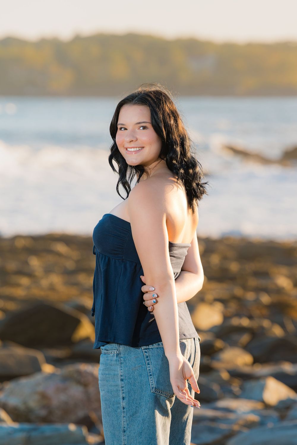 Someone in a navy top and jeans poses casually by a rocky beach shoreline at golden hour.