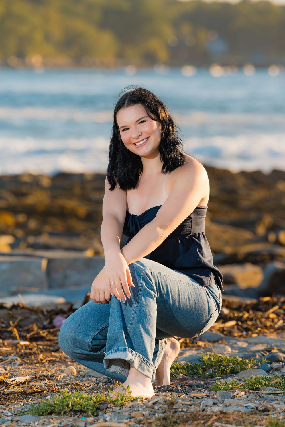 Person in blue jeans and dark top posing casually by the beach at golden hour with water and rocky shore in background.