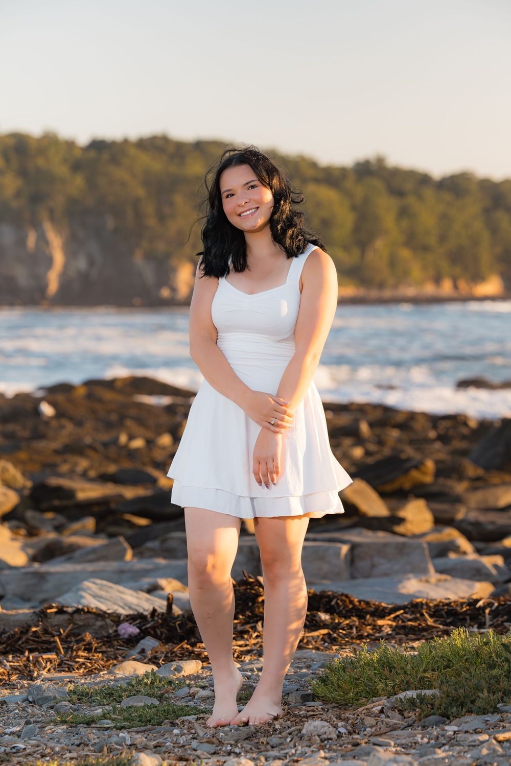 A white-dressed figure poses along a rocky beach shoreline at sunset with ocean waves in the background.