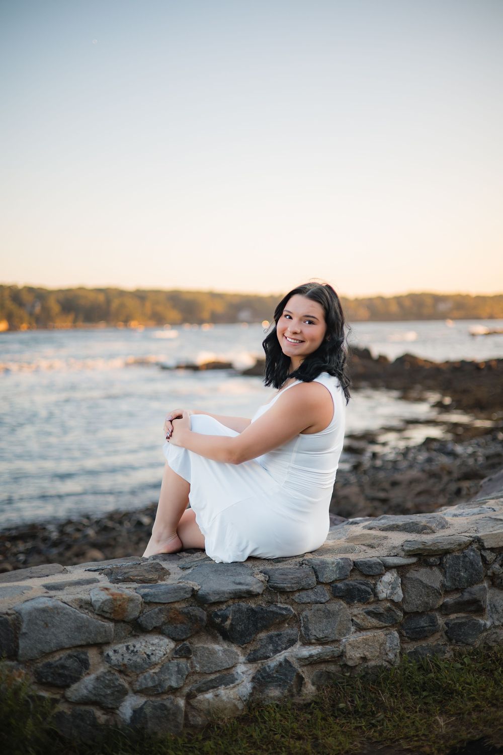 A figure in white reclines on rocky coastal outcrops near calm ocean waters at dusk.