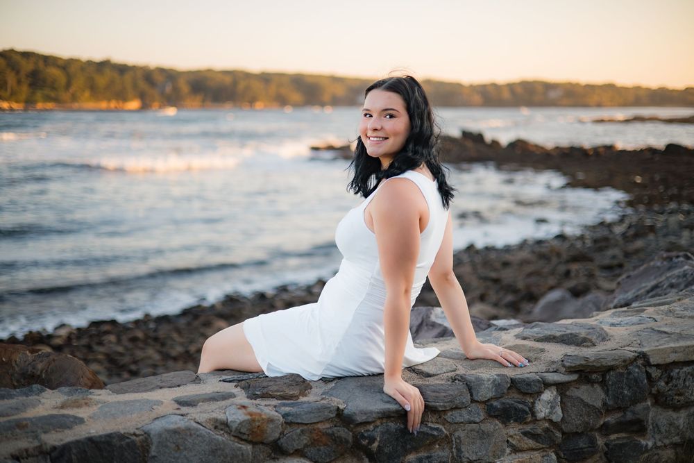 A figure in a white dress poses gracefully on rocky coastal cliffs at sunset with ocean waves in the background.