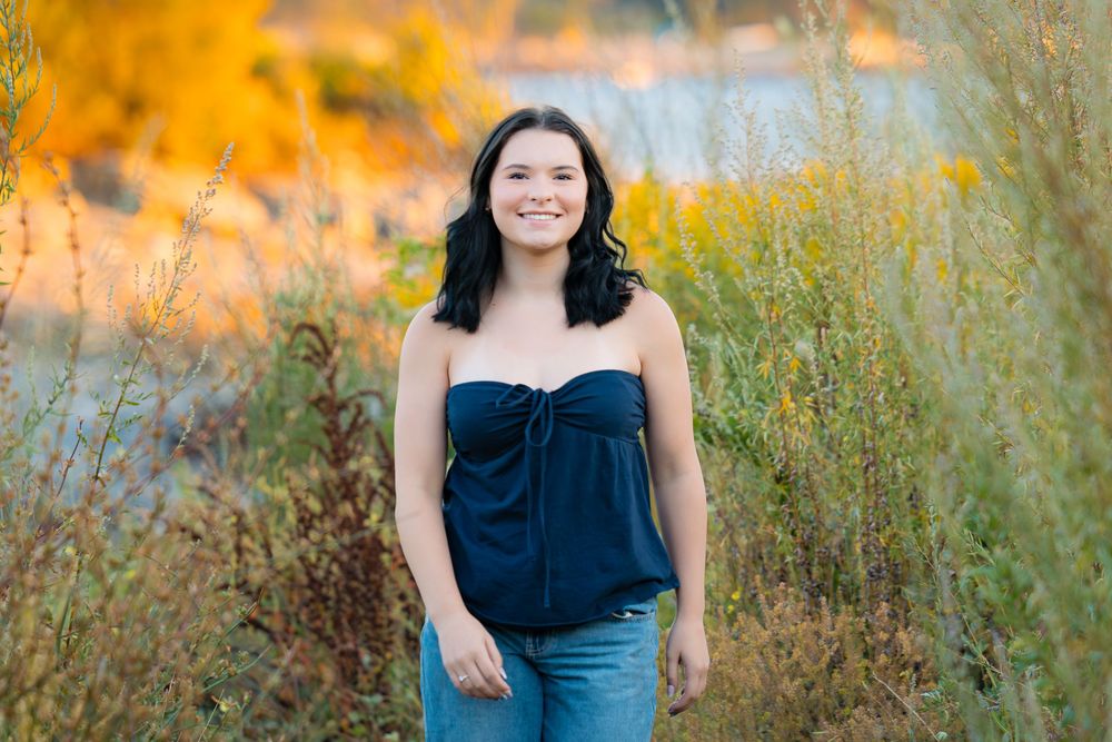 Woman in navy blue dress poses outdoors during golden sunset among tall grass and wildflowers.