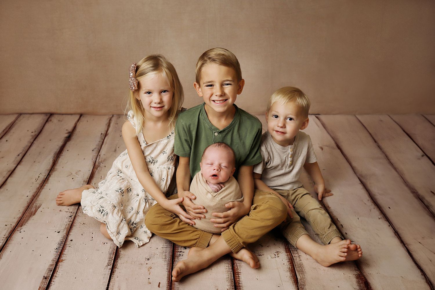 Four children sit together on a wooden floor in a studio setting wearing coordinating neutral colored outfits. West Jordan newborn photos