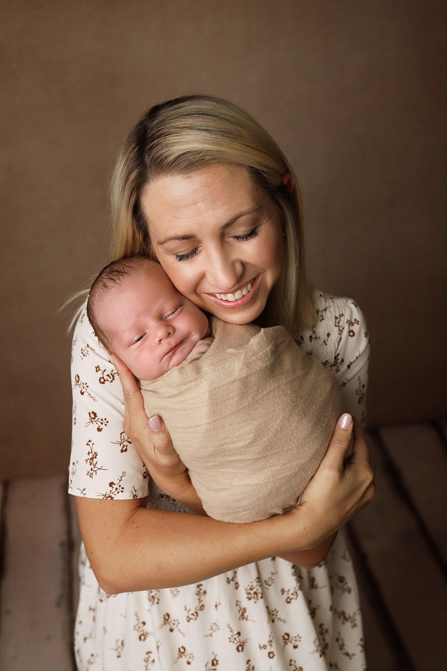 A tender moment captured as someone in a floral dress cradles a sleeping newborn baby wrapped in beige fabric.