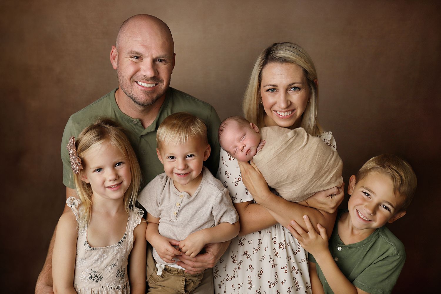 A warm family portrait of six people including children in neutral tones against a brown backdrop.