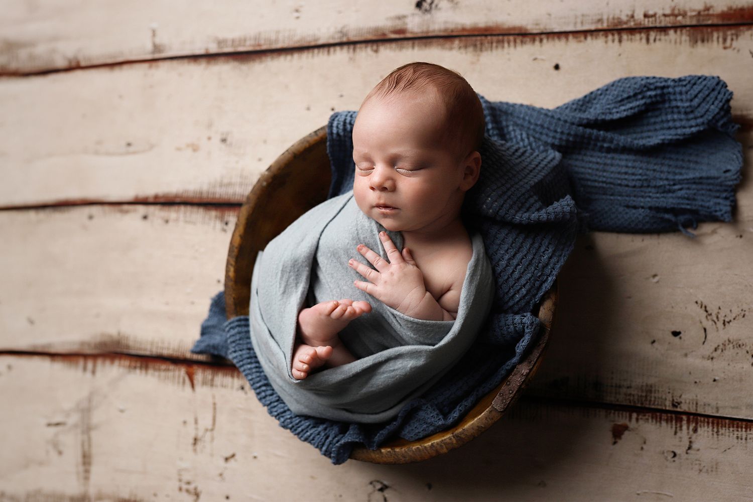 A sleeping newborn baby wrapped in soft gray and navy blue swaddle blankets on a white wooden background.