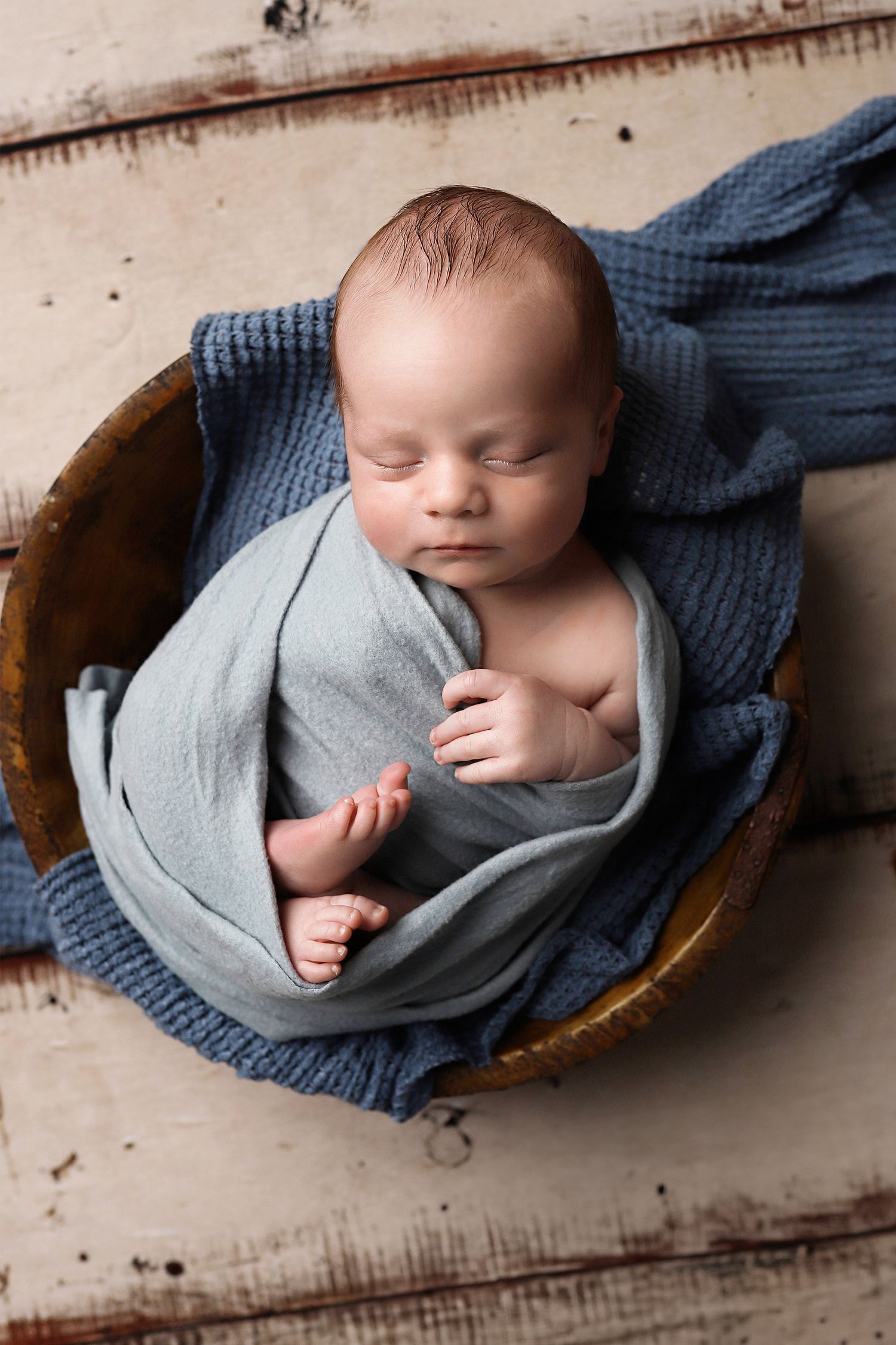 Sleeping newborn wrapped in gray and blue knit blankets lying in rustic wooden bowl in professional photography setup. Utah newborn photos