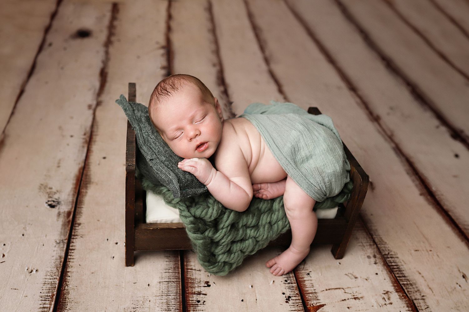 Sleeping newborn on tiny wooden bed with green blanket resting on rustic wooden floor.