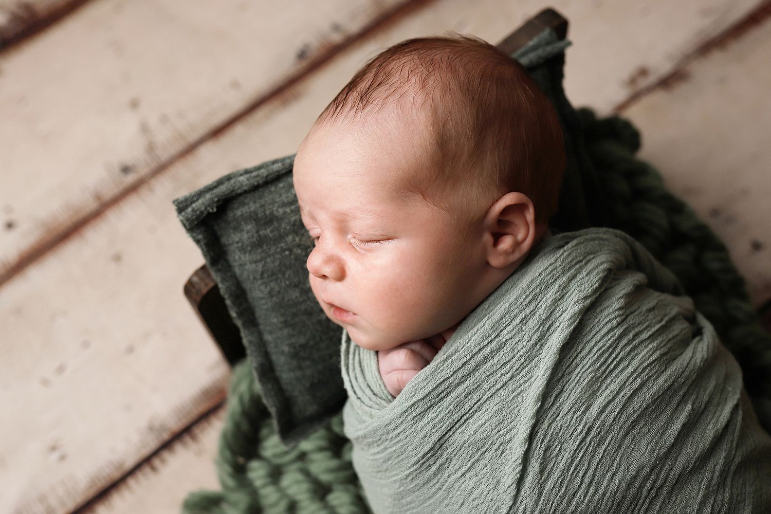 Peaceful sleeping newborn wrapped in soft sage green swaddle blanket on wooden backdrop.
