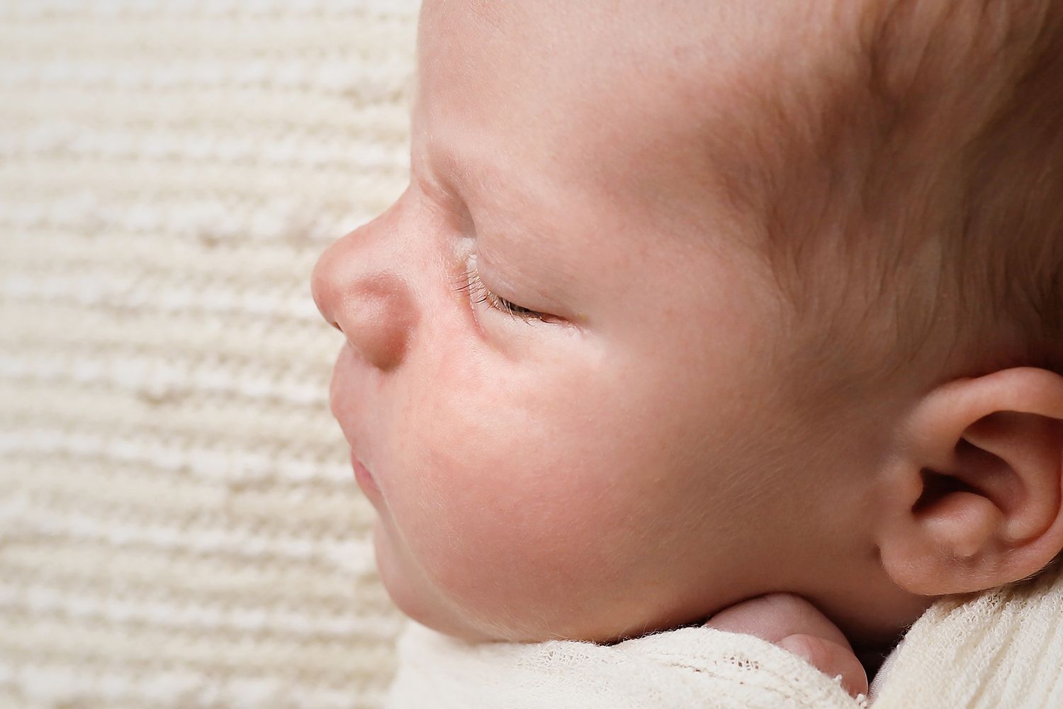 A closeup profile view of a sleeping infant with rosy cheeks resting on white fabric.