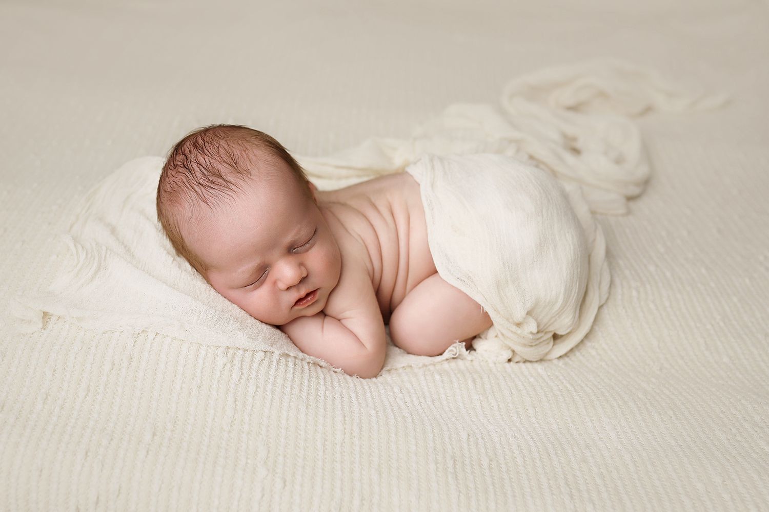 Sleeping newborn baby swaddled in white fabric rests peacefully on textured cream-colored blanket. Utah County newborn photographer.