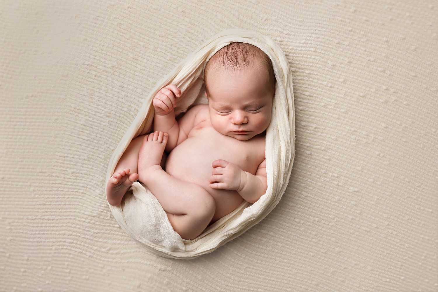 Sleeping newborn baby wrapped in soft white fabric on textured beige backdrop during professional portrait session.