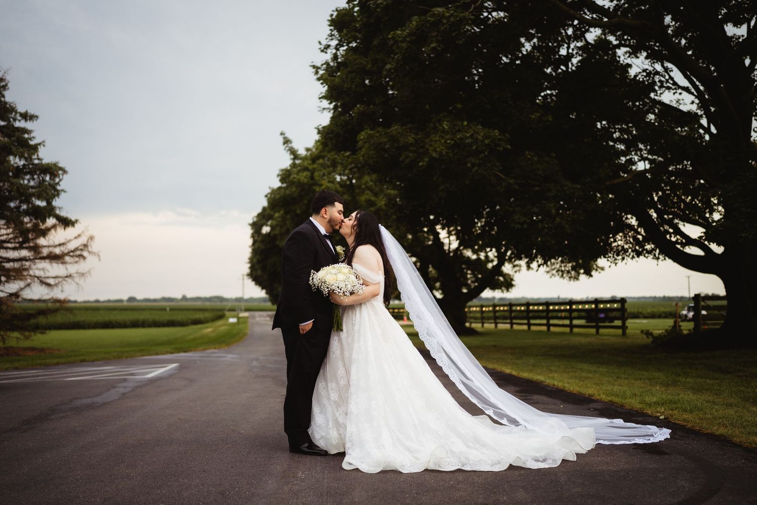 A couple in wedding attire embraces on a country road with a long flowing veil and tree backdrop at sunset.