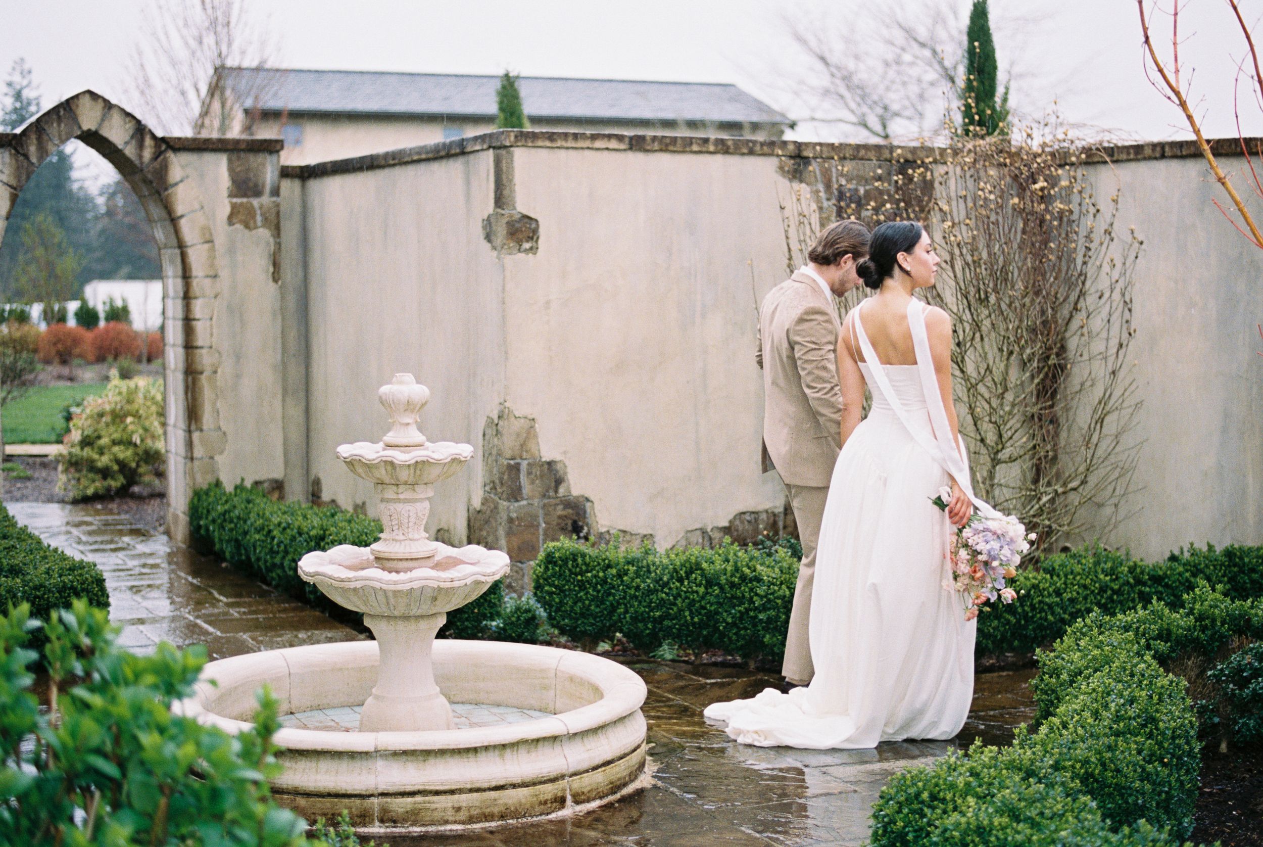 A person in a white wedding dress poses gracefully next to an elegant outdoor fountain in a garden setting.