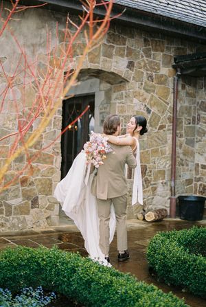 A romantic wedding moment captured at a rustic stone chapel with blooming coral branches and manicured boxwood gardens.