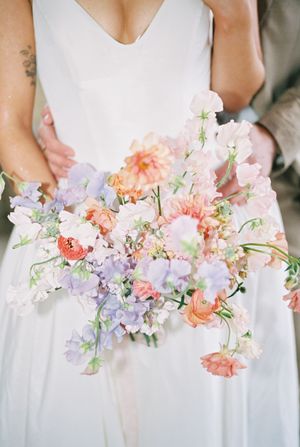 A row of elegant wedding bouquets featuring coral peonies and purple sweet peas held against white dresses.